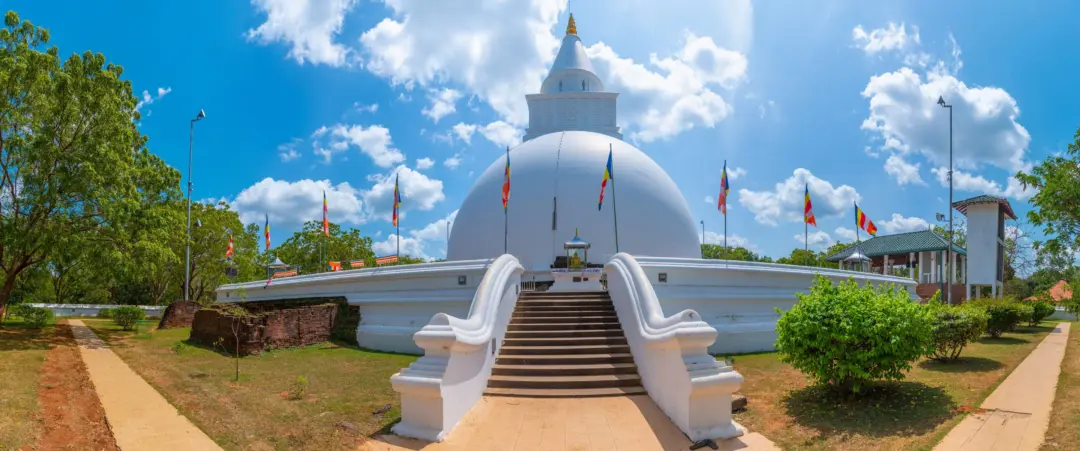 White Buddhist stupa with a large central dome, towering steps, and colorful flags under a bright blue sky.