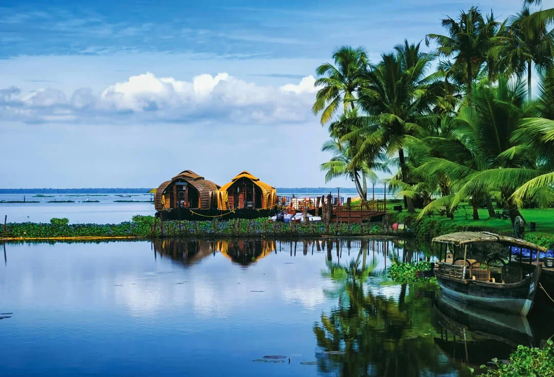 Two thatched-roof houseboats on a calm waterway, with palm trees and a docked boat, reflected in the still water.