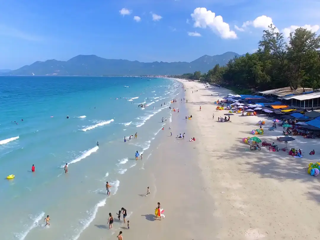 Wide beach with turquoise water, light sand, and families under colorful umbrellas along the shore.