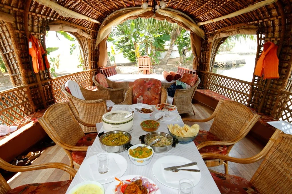 Dining table set for a meal inside a thatched riverboat, surrounded by wicker chairs and tropical greenery. -OR-