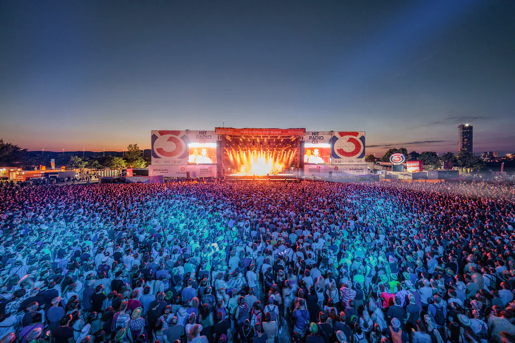 Huge outdoor concert crowd faces a brightly lit stage at dusk with banners and screens on the sides of the stage, creating a festival atmosphere.