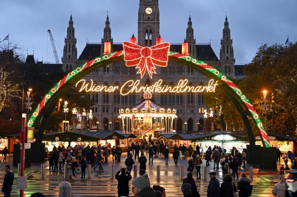Wiener Christkindlmarkt illuminated arch with a red bow over a busy Christmas market in front of a historic building at dusk/night.