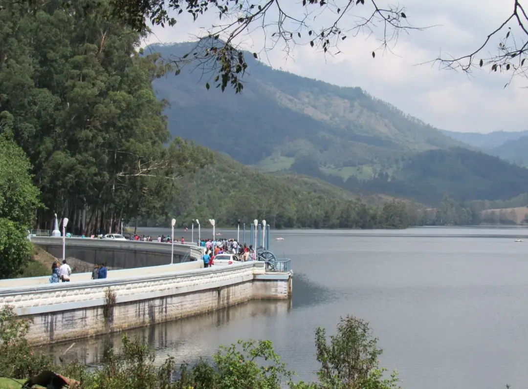 People walking along a curved lakeside promenade with a calm lake and forested hills in the background.