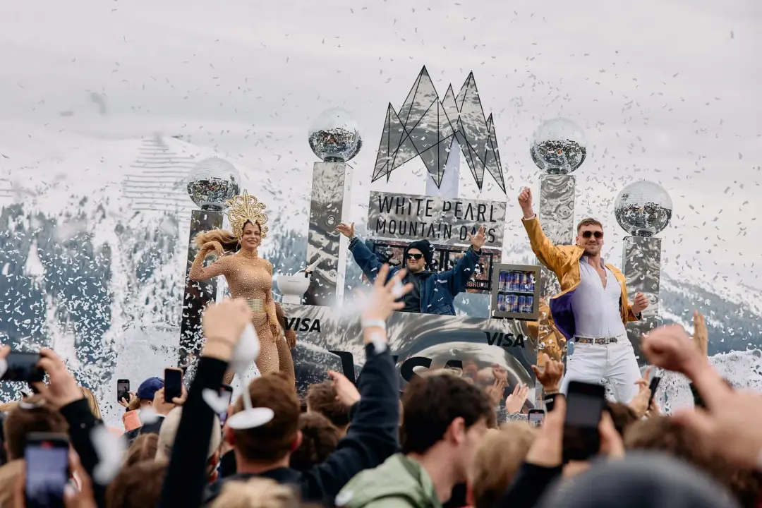 Performers on a festival float pose and celebrate as confetti rains over a cheering crowd in a snowy mountain setting.