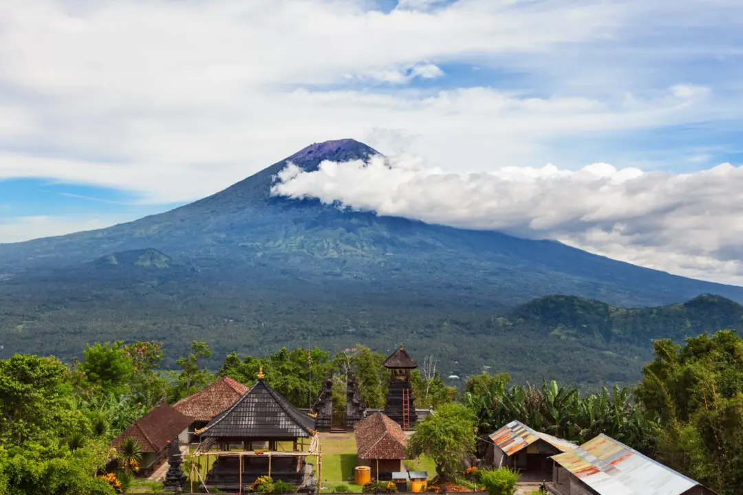 Conical volcano rising above a green valley with a small tropical village in the foreground, thatched-roof huts visible under blue skies.