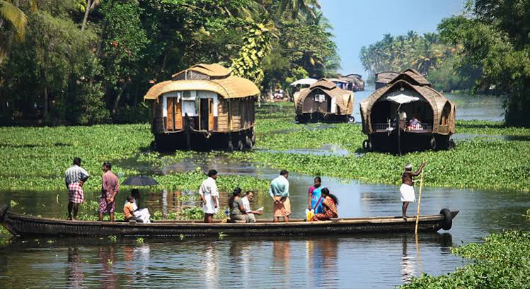 Group of people riding a long wooden boat on a calm river with thatched-roof houseboats in the lush, green shoreline behind them.