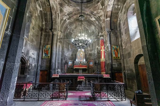 Interior of a stone church chapel with an ornate altar, arched ceiling, hanging chandelier, and red banners on either side.