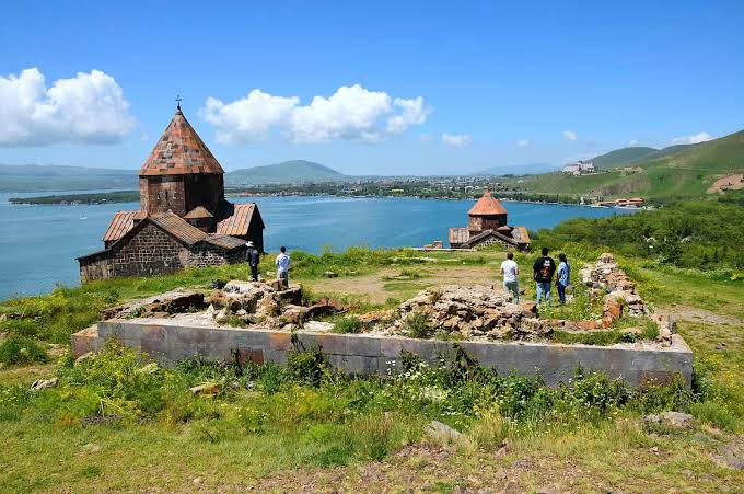 Ruined stone fortress with conical towers on a grassy hill overlooking a calm blue bay, with a few visitors exploring the site.