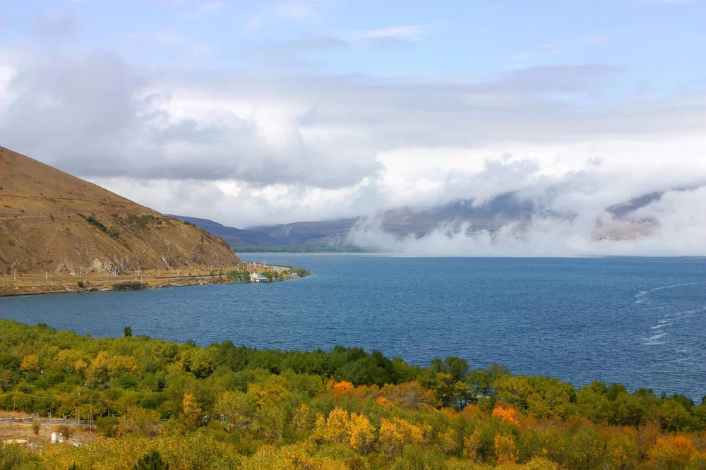 Autumnal lakeside landscape: colorful trees in the foreground, a blue lake, and hills with clouds in the distance.