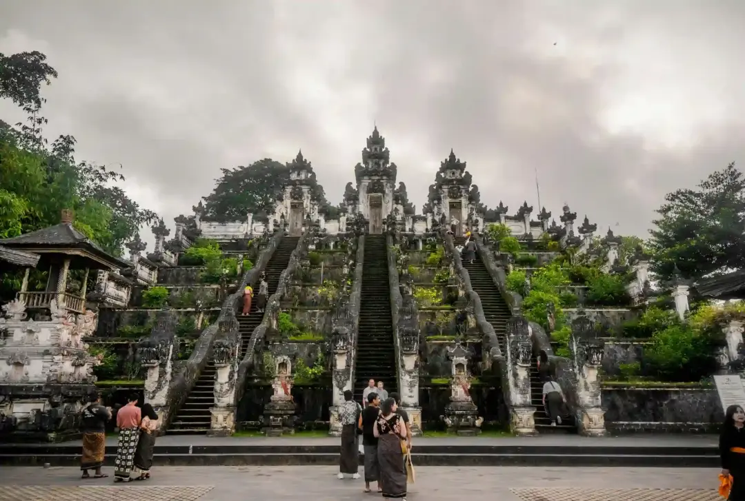 Ancient multi-tiered stone temple with central and side staircases, surrounded by greenery and visitors in traditional attire at the base.