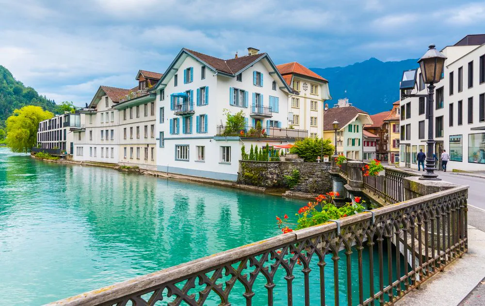 Colorful houses line a turquoise river with a decorative railing in the foreground and mountains in the distance.