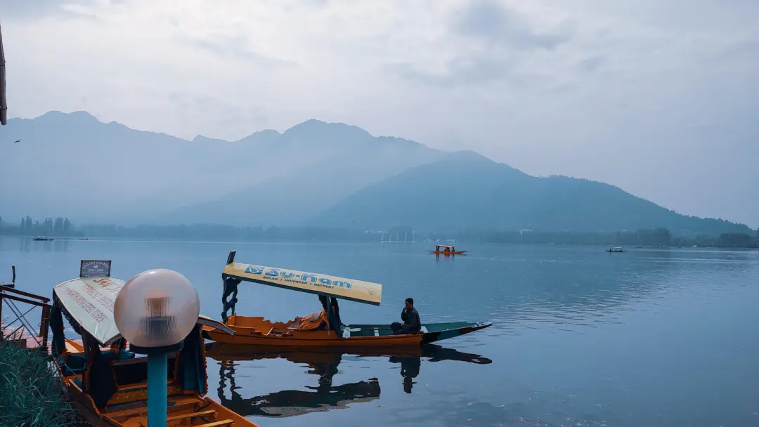 Calm lake with orange boats docked near the shore, misty mountains in the distance.