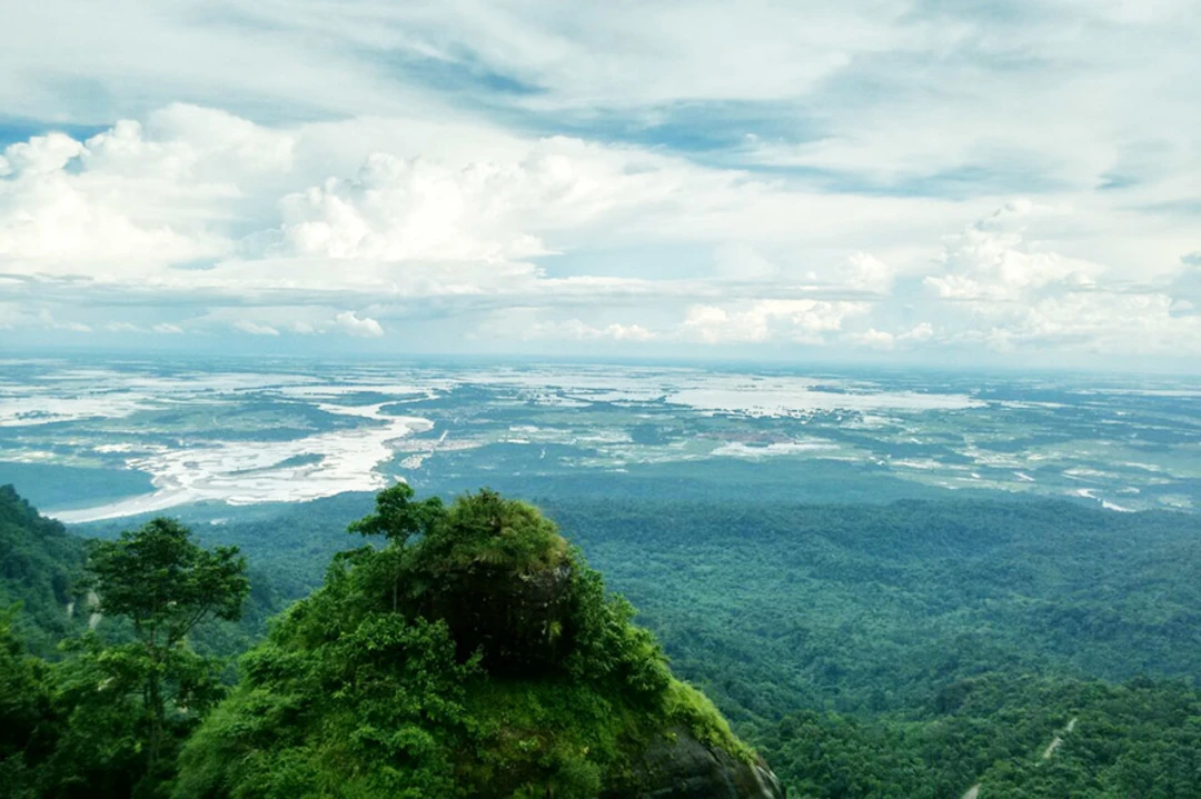 Green mountainside viewpoint overlooking a broad river valley and scattered wetlands under a bright, cloudy sky.