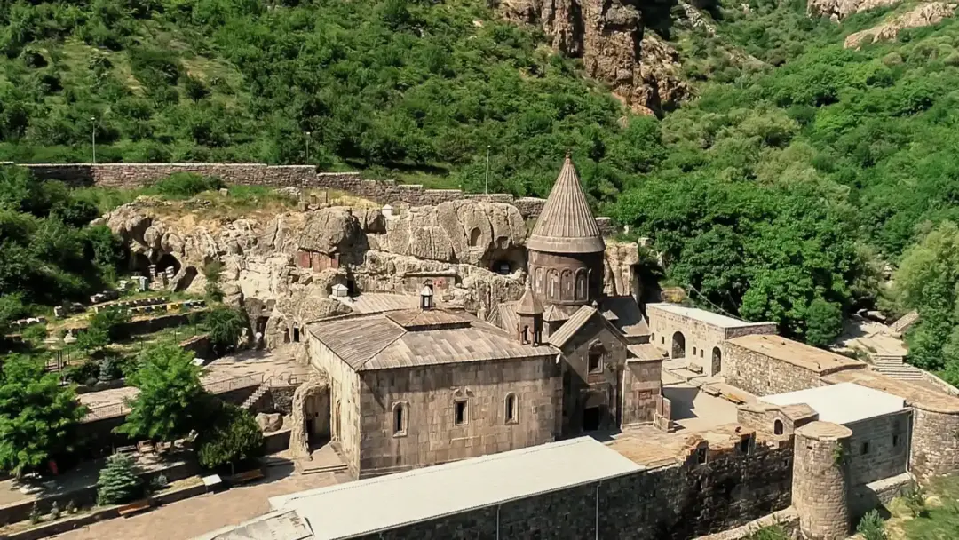 A medieval stone church with a tall conical roof in a fortress-like complex set on a rocky hillside, surrounded by trees.