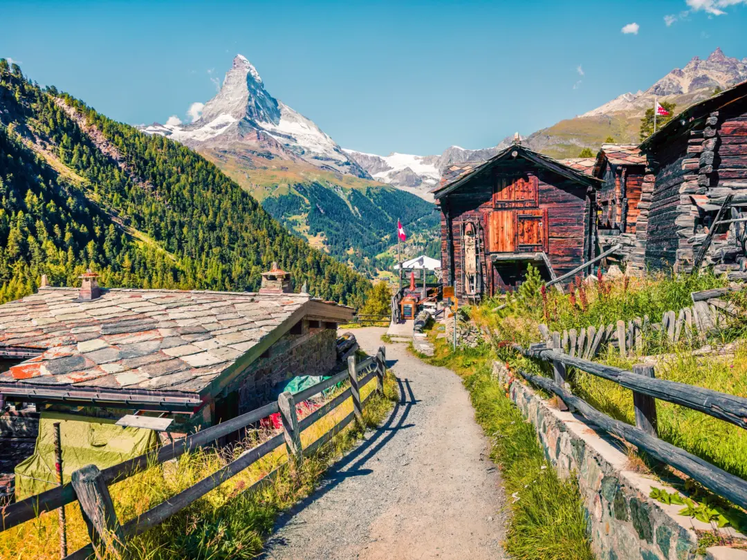 Path through a rustic mountain village with wooden chalets and stone fences against a backdrop of green hills and the Matterhorn.